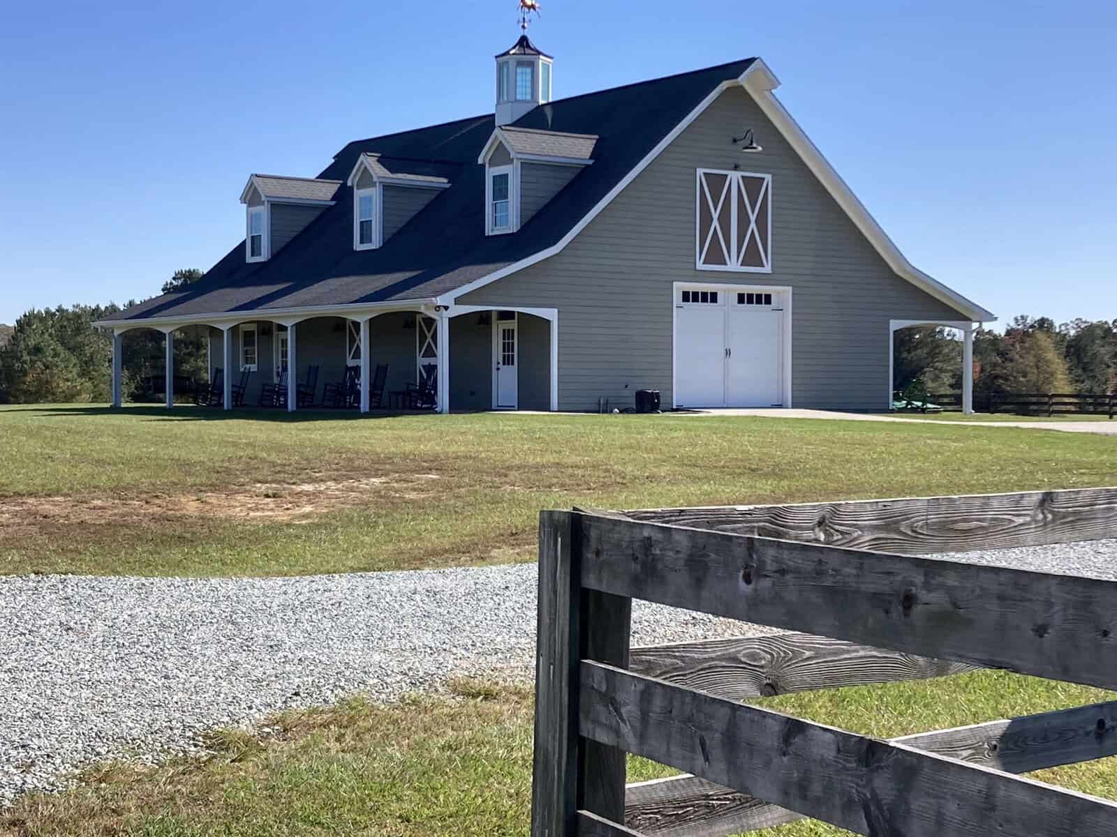 A large barn with a gray exterior and a gambrel roof sits on a grassy field under a clear blue sky. The barn features white doors and dormer windows. A gravel path leads up to the barn, and a wooden fence is in the foreground.