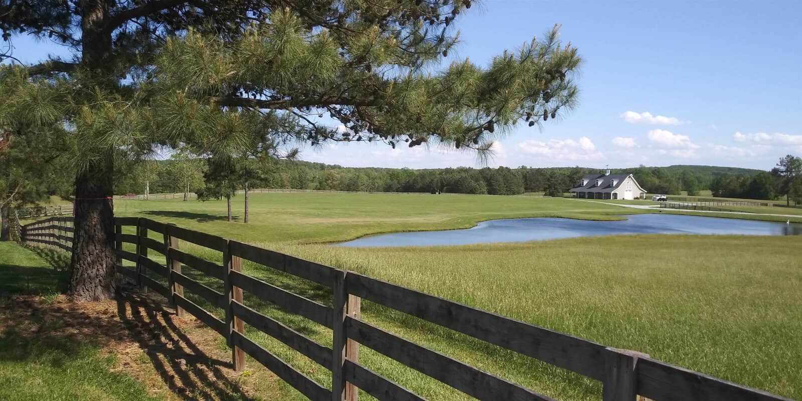 Experience the serene charm of Rougemont with a scenic view at 4708 Uzzle Road. A wooden fence frames the lush green pasture, while a tranquil pond and a distant white building add to the picturesque setting. A tree casts gentle shade on this sunny day under the clear blue sky.