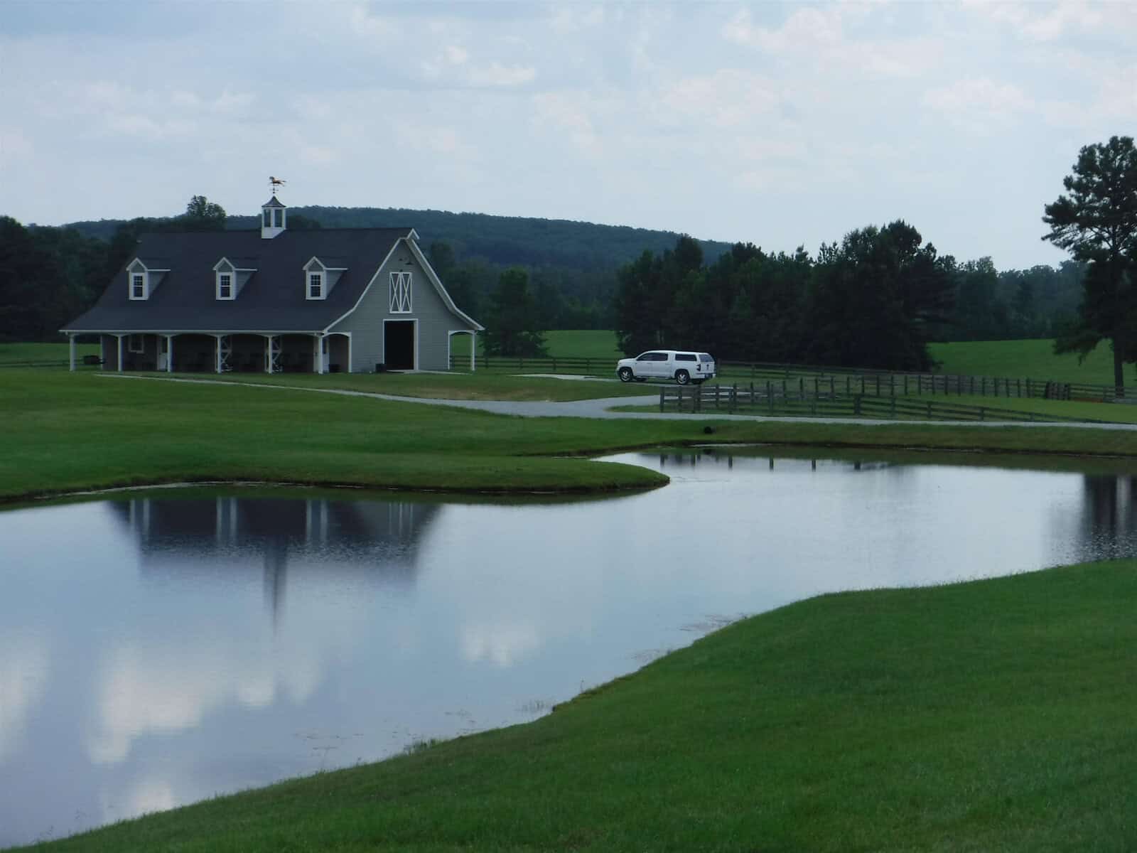 A scenic landscape featuring a large grassy field with a pond in the foreground. In the background, there's a barn-style building with a weather vane on top and a white SUV parked nearby. Trees and hills are visible under a cloudy sky.