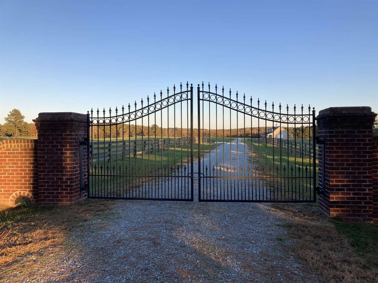 A black wrought iron gate set between two red brick pillars, leading to a gravel path that stretches into the distance. The path is lined with fences and grass, under a clear blue sky.