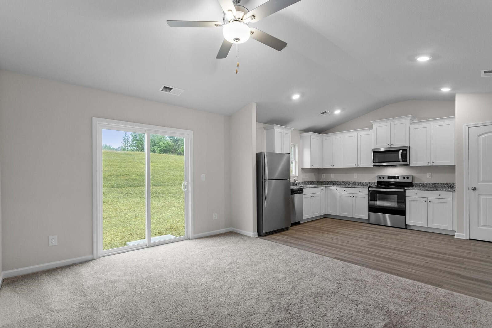 A modern, spacious kitchen and living area with white cabinets, stainless steel appliances, and wood flooring. A ceiling fan is on the white ceiling. A sliding glass door opens to a grassy outdoor area, viewed from a carpeted living space.