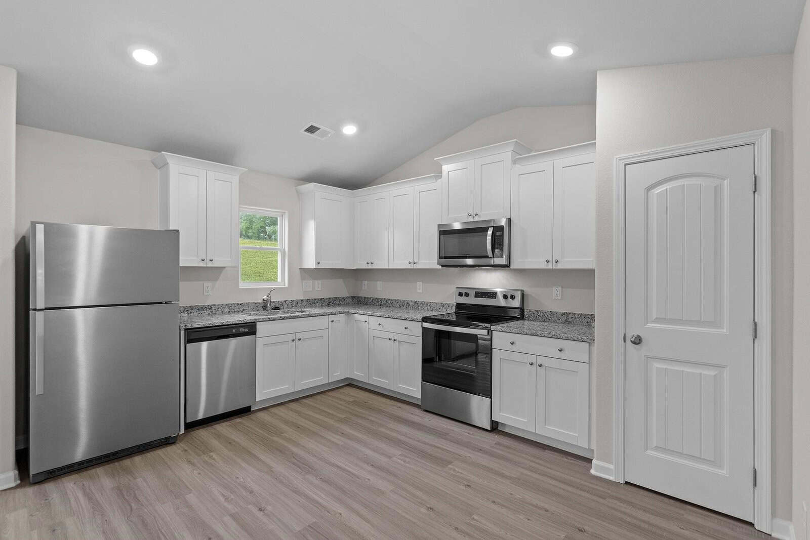 A modern kitchen with white cabinets, stainless steel appliances, and a gray granite countertop. It features a fridge, dishwasher, oven, and microwave. The room has recessed lighting and wood flooring, with a small window providing natural light.