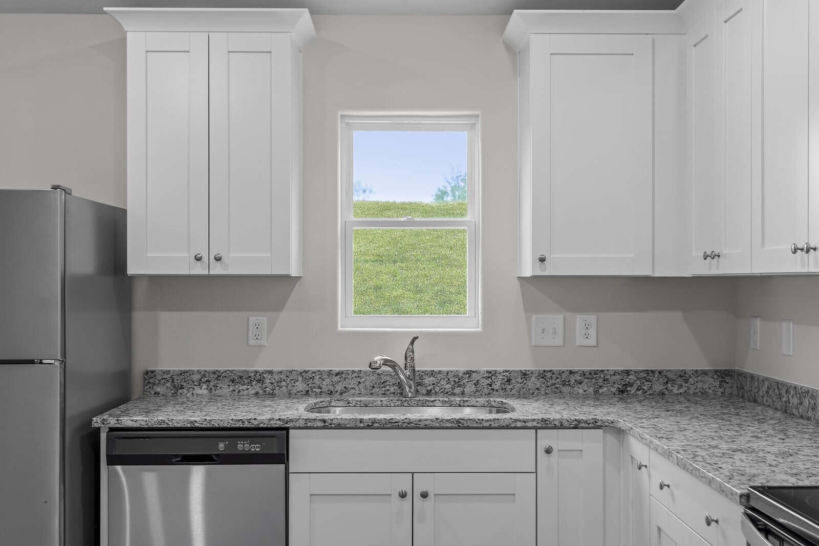 A modern kitchen featuring a granite countertop, stainless steel appliances including a refrigerator and dishwasher, white cabinets, and a central window displaying a grassy outdoor view.