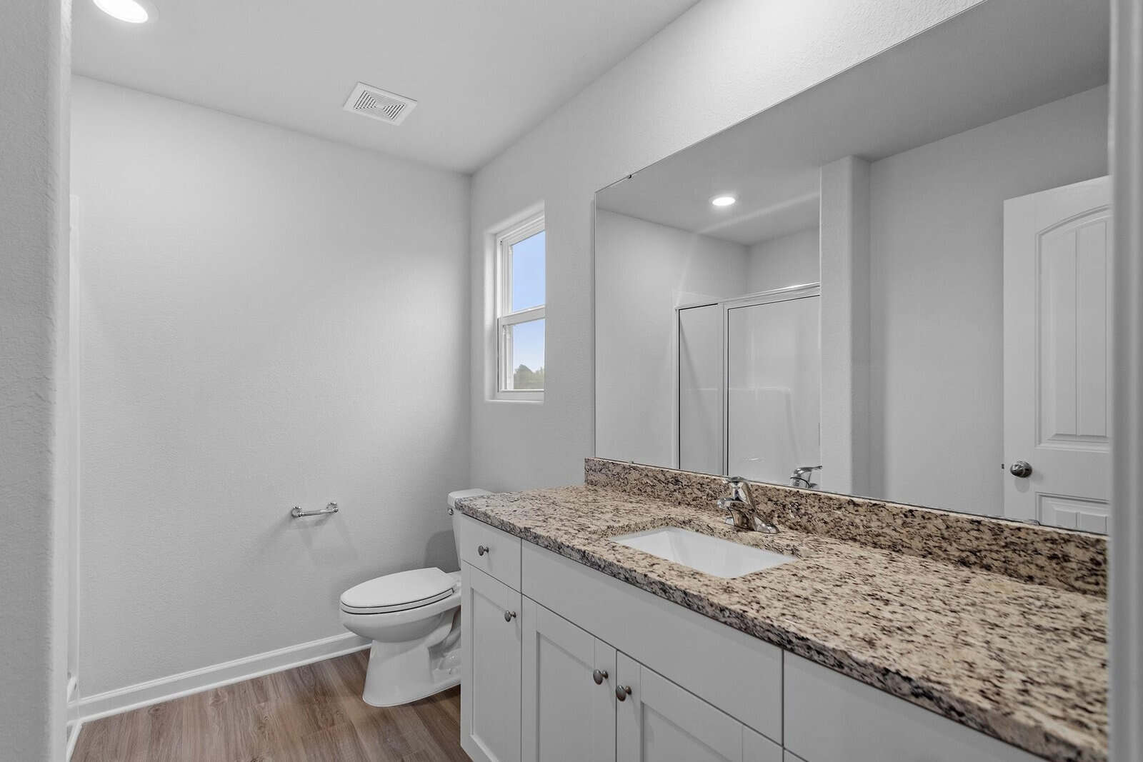 A modern bathroom with a granite countertop and white cabinets under a large mirror. The room features a toilet, a shower with a glass door, wood flooring, and a small window letting in natural light.