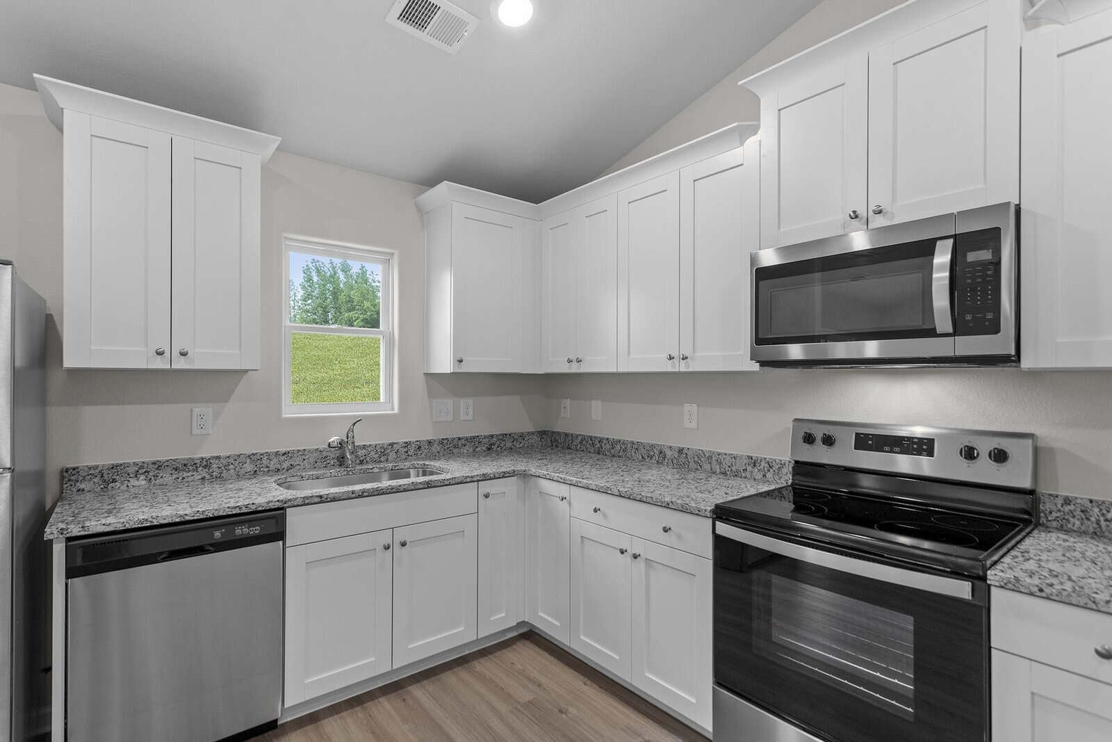A modern kitchen with white cabinets, granite countertops, and stainless steel appliances, including a stove, microwave, and dishwasher. A small window lets in natural light, illuminating the light hardwood floor.