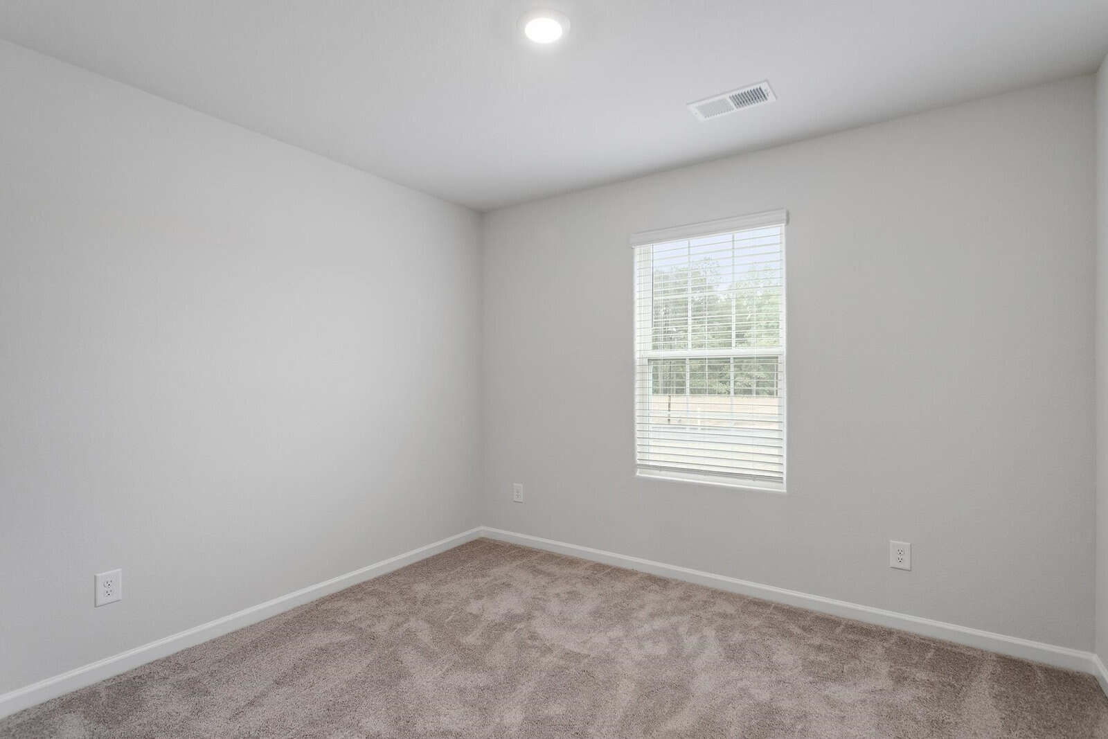 A small, empty room with light gray walls and beige carpet. A window with blinds is on one wall, allowing natural light to enter. The ceiling has a recessed light fixture.