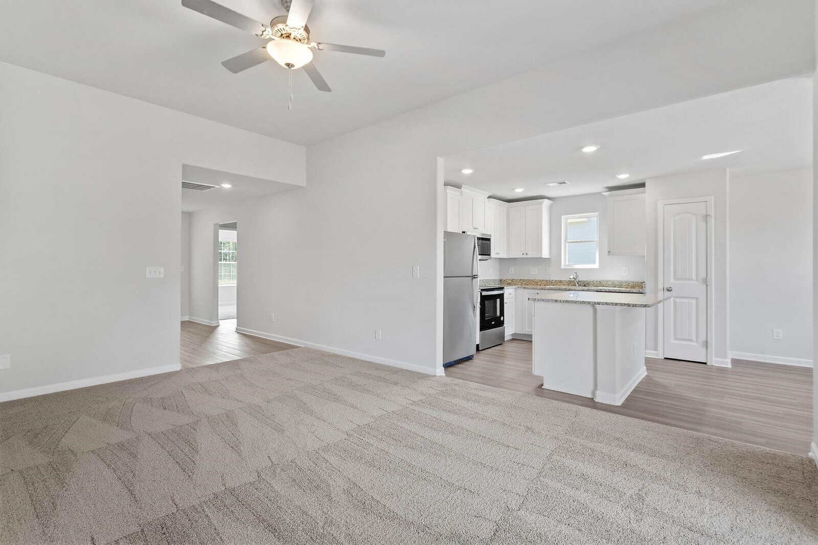 Spacious open-concept living area with beige carpet and ceiling fan. Adjacent to a modern kitchen featuring white cabinets, stainless steel appliances, and hardwood floors. Bright and airy with light pouring through a window and a door.