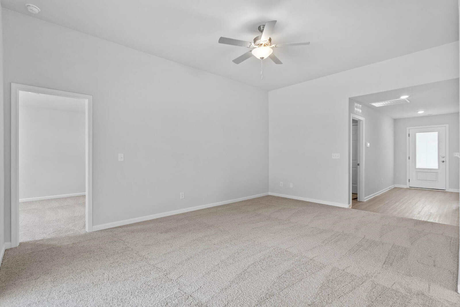 A bright, empty room with light gray walls and beige carpet. A ceiling fan is centered on the ceiling. There are three doorways: one leading to another room and two leading to a hallway with a wooden floor and a distant white door.