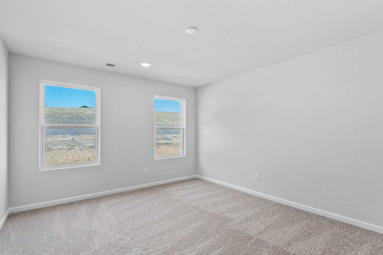 An empty room with light gray walls and carpeted flooring. Two large windows on one side illuminate the space with natural light. The view outside shows a clear sky and a grassy hill.
