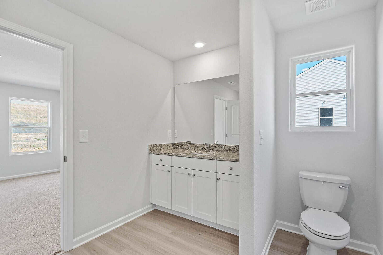 A bright bathroom with light wood flooring, featuring a white vanity with a granite countertop, a large mirror, and a toilet. A small window brings in natural light. The room connects to a partially visible adjacent carpeted room.