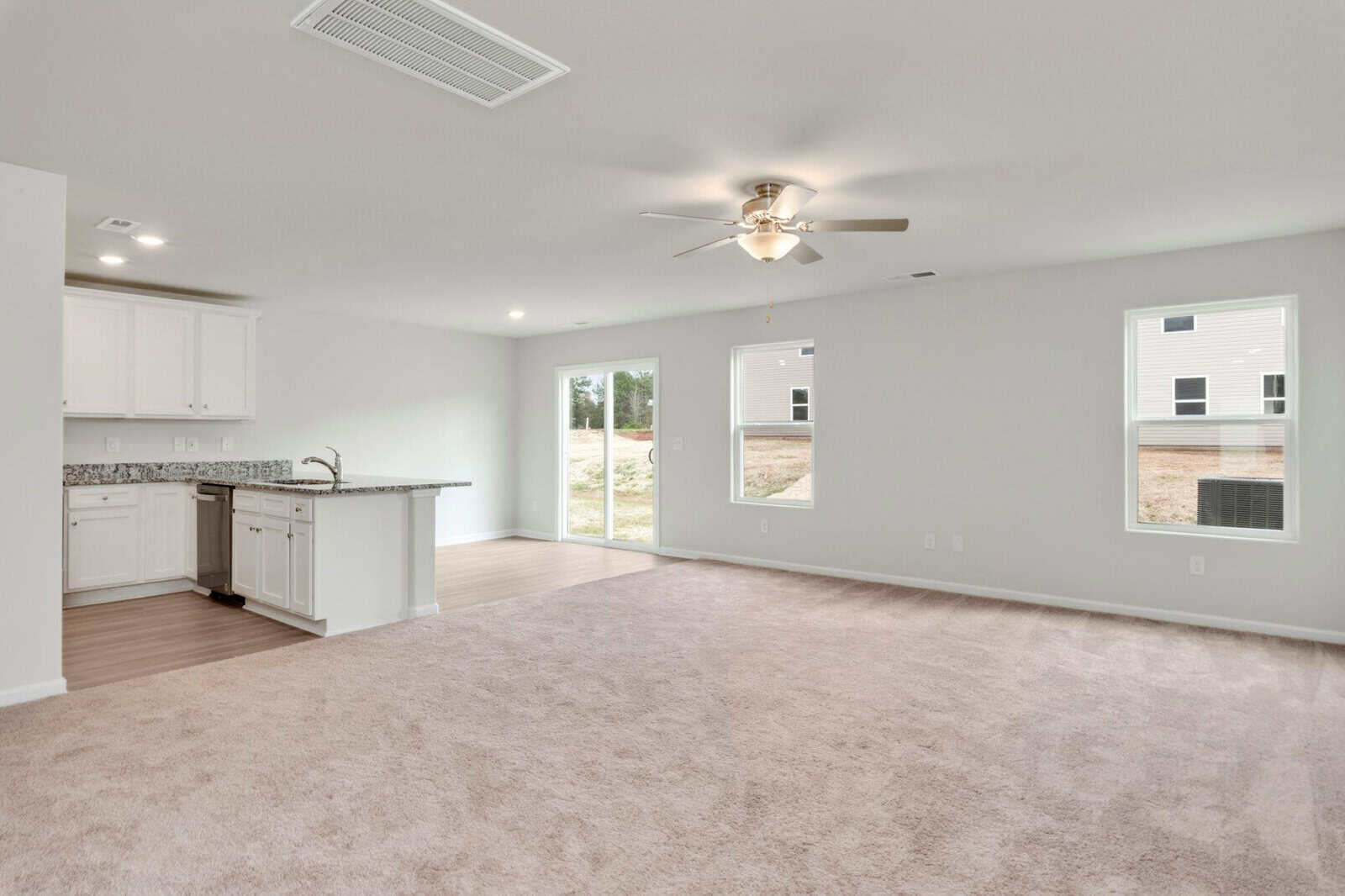 Spacious living area with beige carpet, adjacent to an open kitchen featuring white cabinets and a granite countertop. Ceiling fan with lights above and large windows offering ample natural light. A sliding glass door leads to the outdoor area.