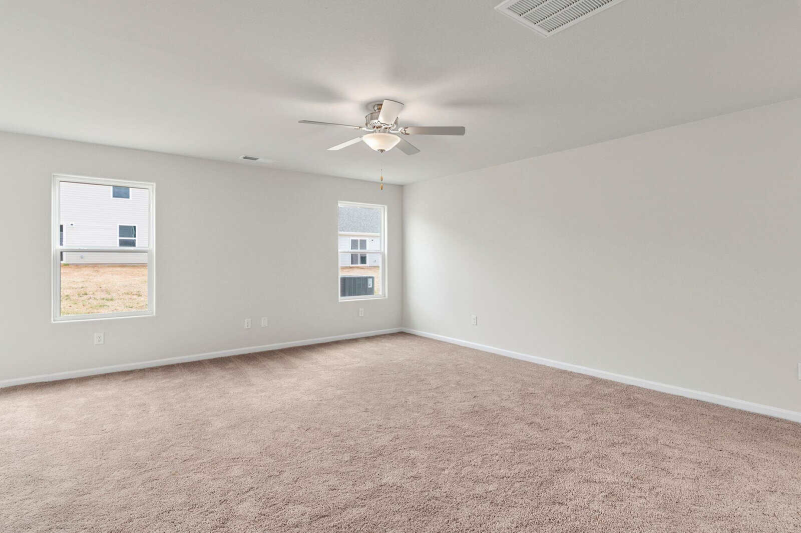 A spacious, empty room with beige carpet and white walls. Two windows let in natural light. A ceiling fan with a light is in the center. The windows offer a view of neighboring houses and a grassy lawn.