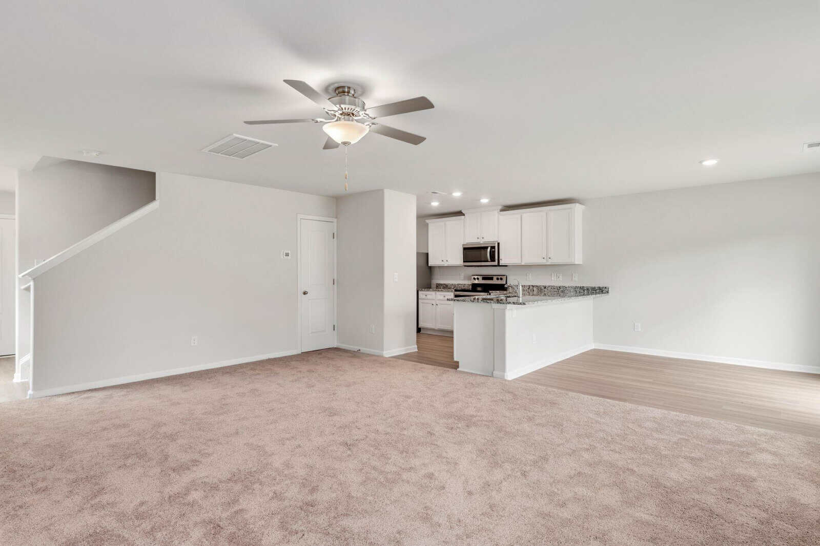 A modern open-plan living space with beige carpet, a ceiling fan, and a kitchen area. The kitchen features white cabinets, a granite countertop, and stainless steel appliances, including a built-in microwave and oven. Stairs lead to an upper level.