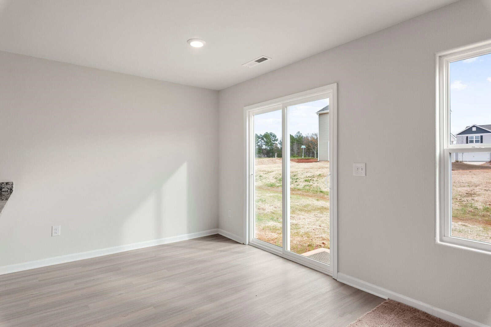 Minimalist empty room with light wood flooring, white walls, and a sliding glass door leading to a backyard. A window on the right lets in natural light.