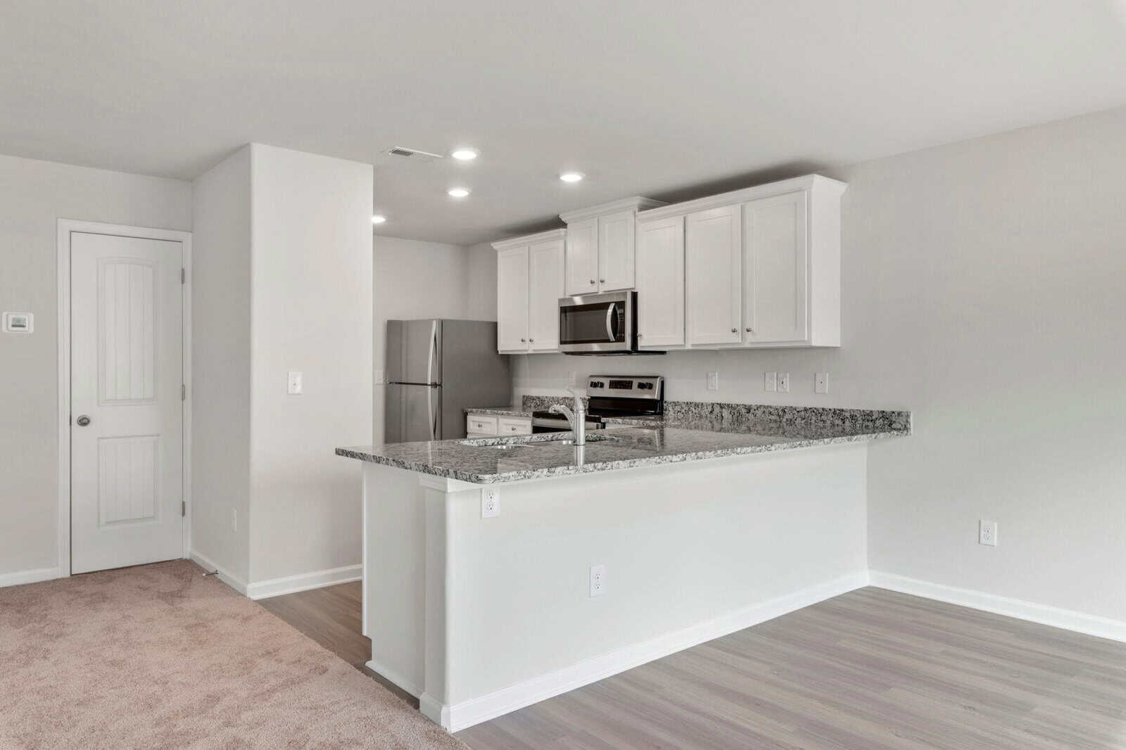 Modern kitchen with stainless steel appliances, white cabinets, a granite countertop, and a bar-style counter with space for seating. The flooring is a mix of carpeted and wood-look surfaces, and recessed lighting illuminates the space.