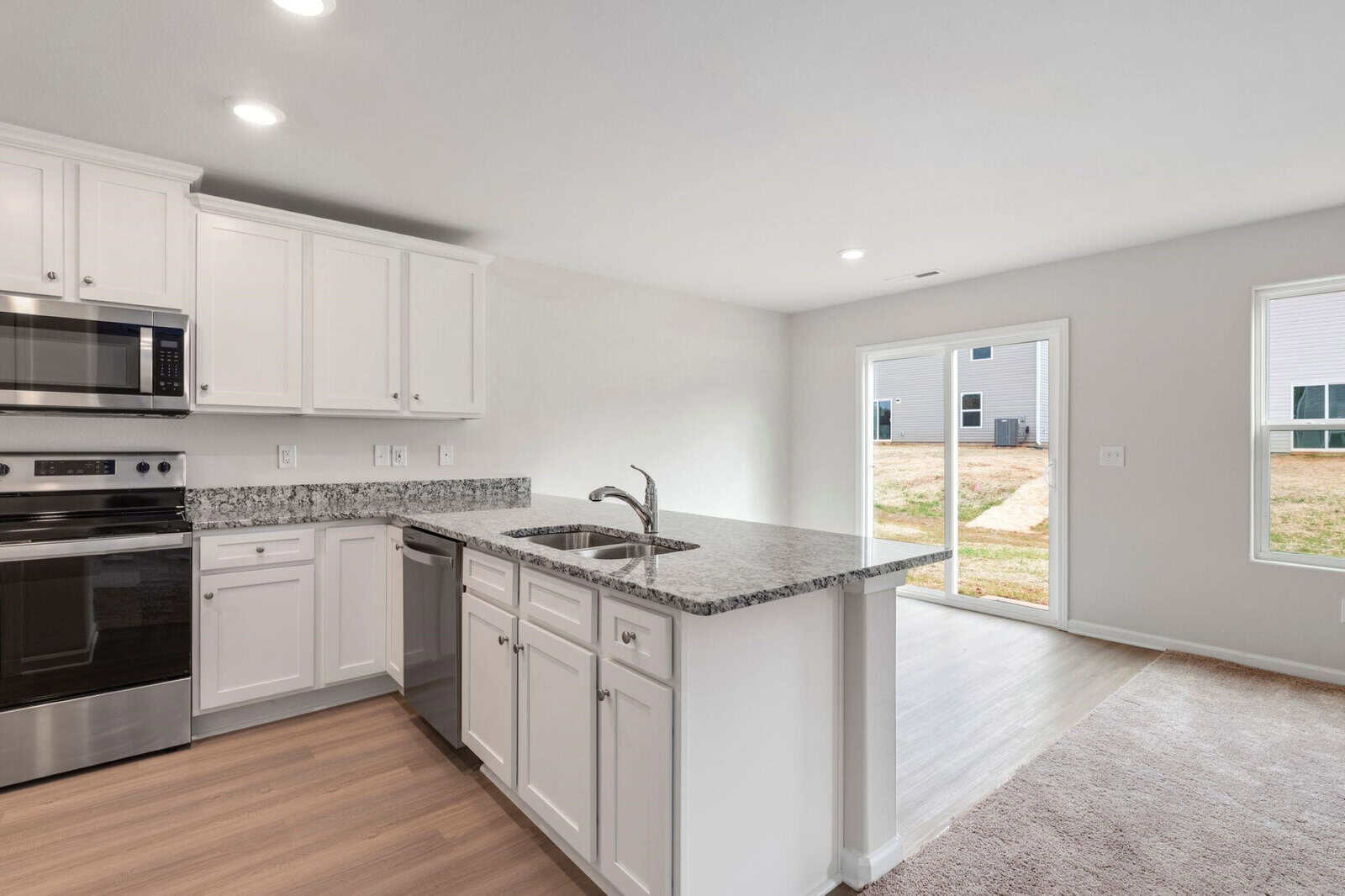 Modern kitchen with white cabinets, stainless steel appliances, and a granite countertop island with a sink. Hardwood flooring transitions to a carpeted area leading to sliding glass doors that open to a backyard with a view of other houses.