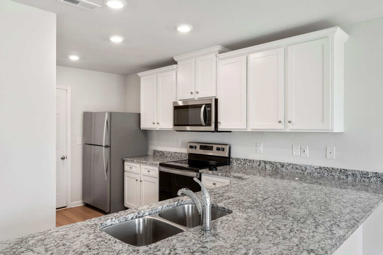 A modern kitchen with white cabinets and granite countertops. Features a stainless steel refrigerator, oven, and microwave. A double sink is built into the counter, and recessed lighting brightens the space.