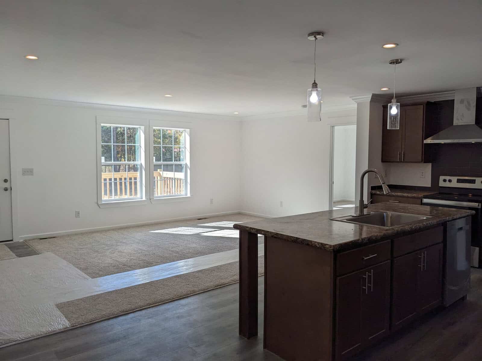 An empty, modern kitchen with hardwood flooring, a central island with a sink, and a view of an adjoining room with a glass door leading to an outside deck.
