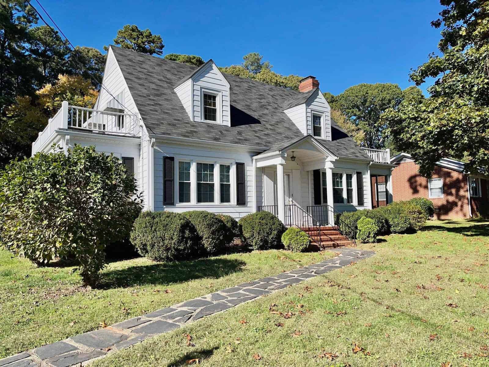 A traditional two-story house with a steep gable roof, dormer windows, and a brick chimney, set against a clear sky with surrounding greenery and a stone pathway leading to the front entrance.