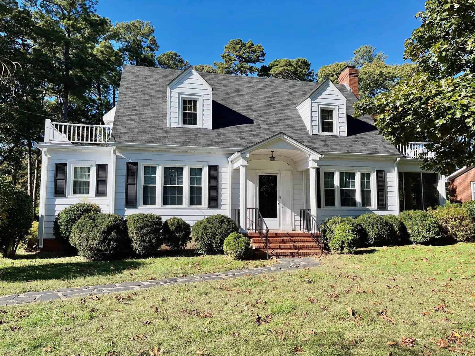 A two-story, cape cod-style house with a gabled roof, dormer windows, and a small front porch surrounded by a green lawn under a clear blue sky.