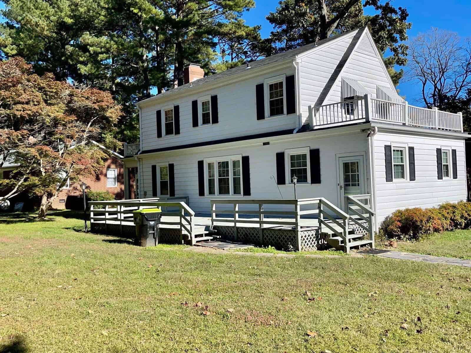 A two-story white house with a balcony and a wooden ramp leading to the front entrance, set against a backdrop of trees and a clear blue sky.