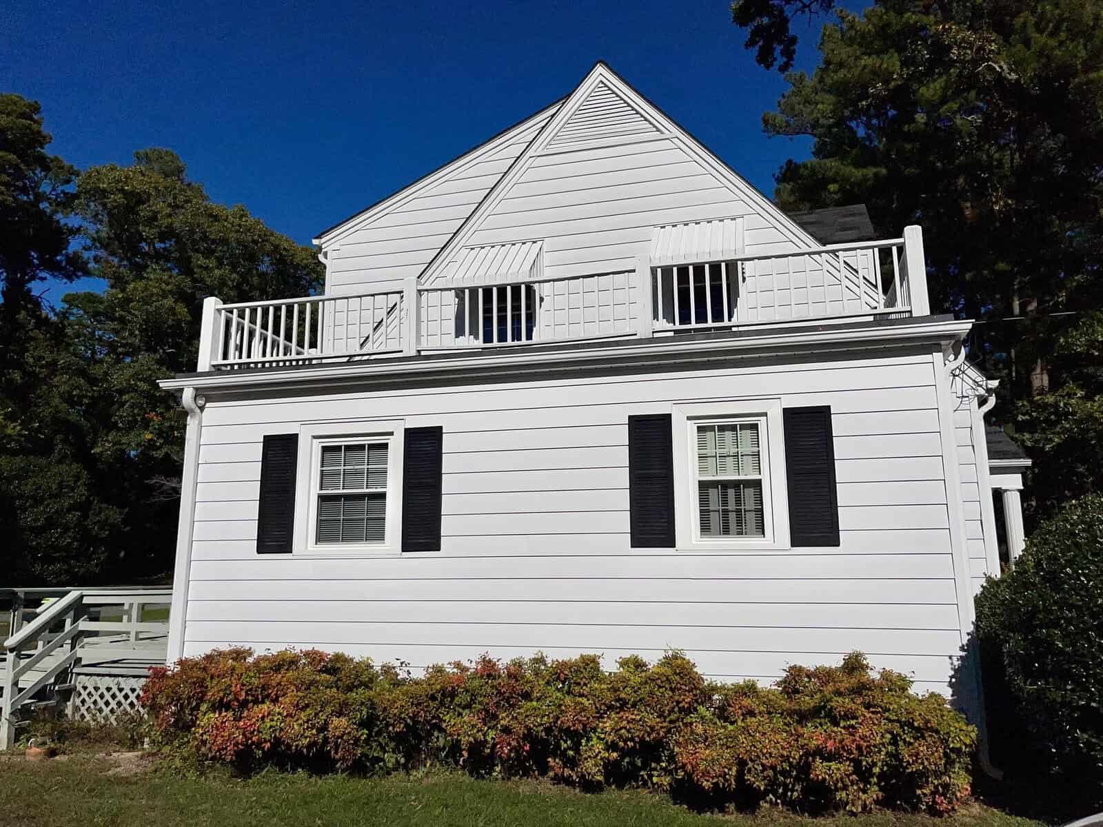 A white two-story house with dark shutters, featuring a second-floor balcony and surrounded by greenery under a clear blue sky.