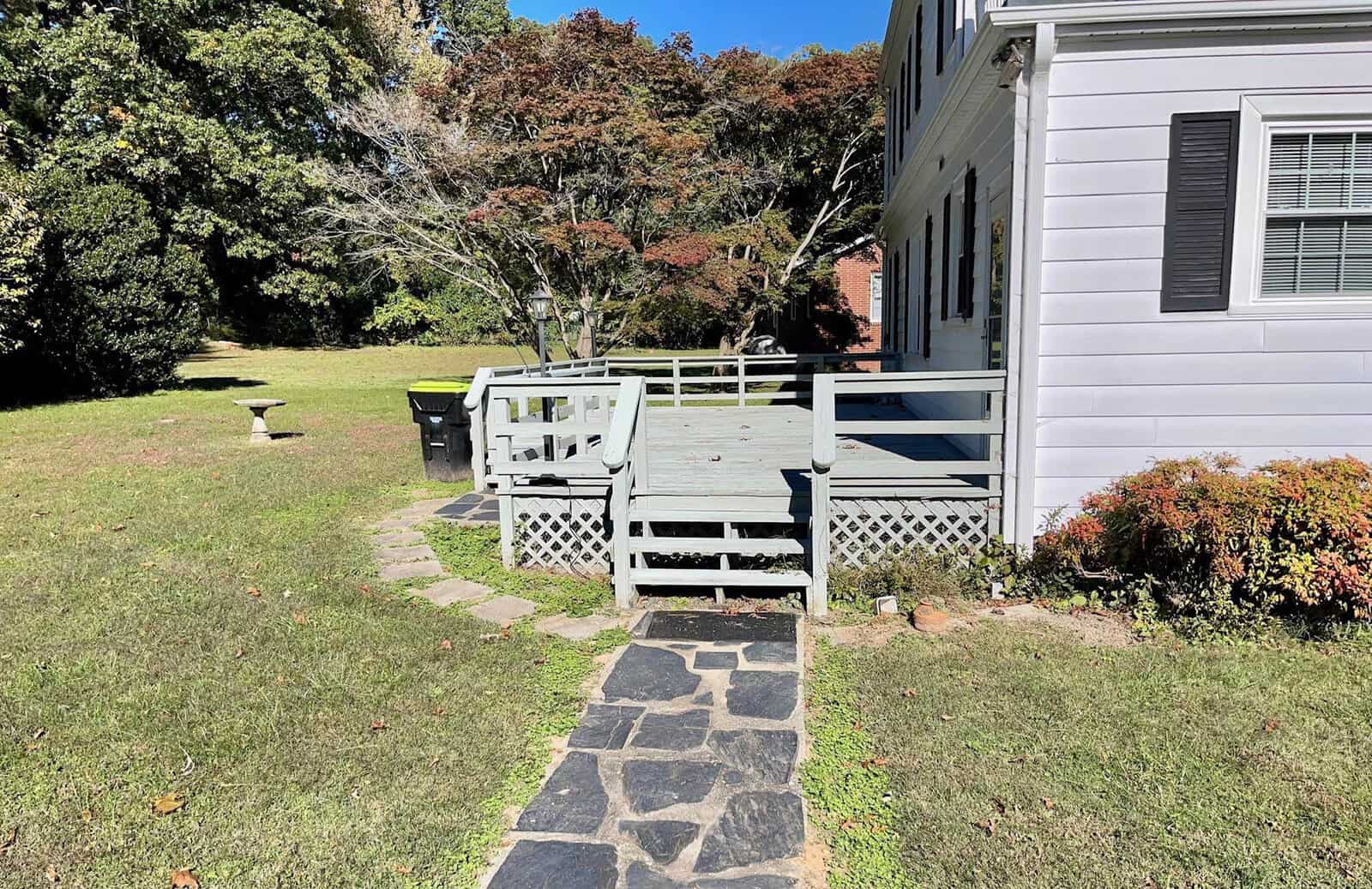 A stone pathway leading to a weathered wooden deck with bench seating next to a white house, surrounded by trees and a lawn.