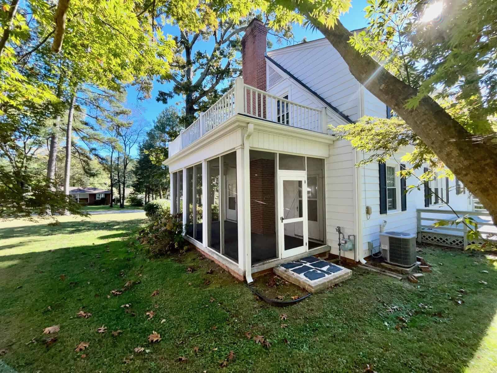 A white house with a screened porch surrounded by lush green trees on a sunny day.