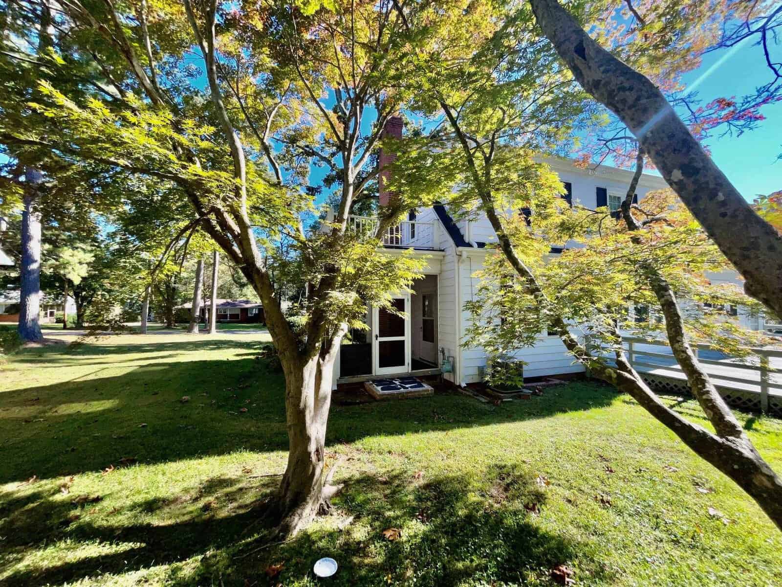 A white two-story house with a porch, partially obscured by leafy green trees on a sunny day with a clear blue sky.