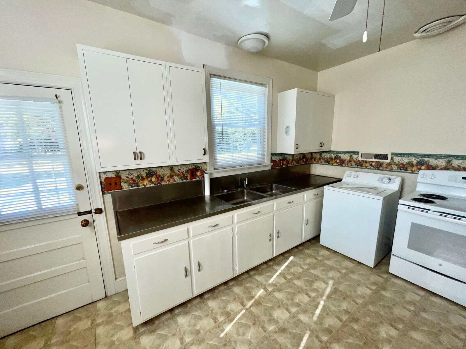 A well-lit kitchen with white cabinetry, a double sink, a washing machine, and a white stove, featuring a window with blinds and a tiled countertop.