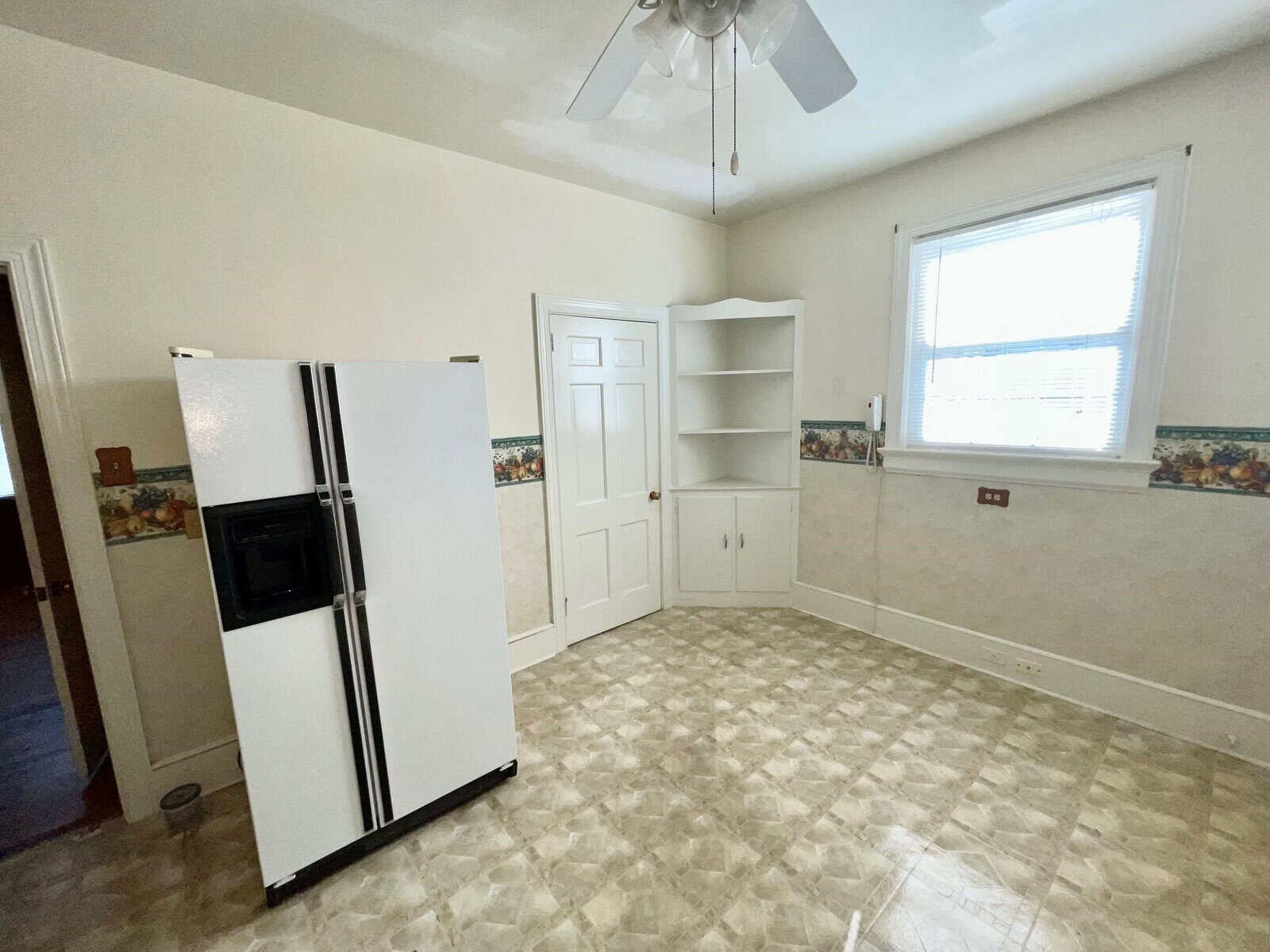 An empty room featuring a white refrigerator on the left, a built-in shelving unit, and a closed white door beside the shelves. there's a window with blinds on the right, a ceiling fan above, and the room has beige walls with a patterned wallpaper border near the ceiling. the floor is covered with vinyl tiles in a diamond pattern.