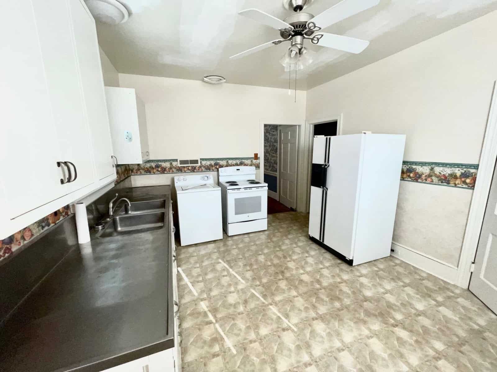A modest kitchen with white appliances, including a refrigerator and stove, with a ceiling fan above and a patterned backsplash.