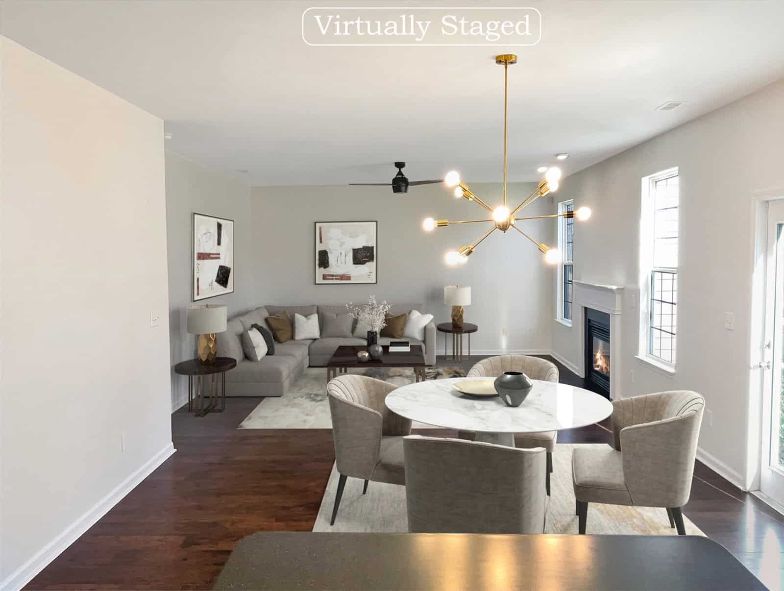 A virtually staged living room with a modern chandelier, round dining table, four upholstered chairs, large beige sectional sofa, and framed art on the walls. The room has hardwood floors and a fireplace with two windows nearby.