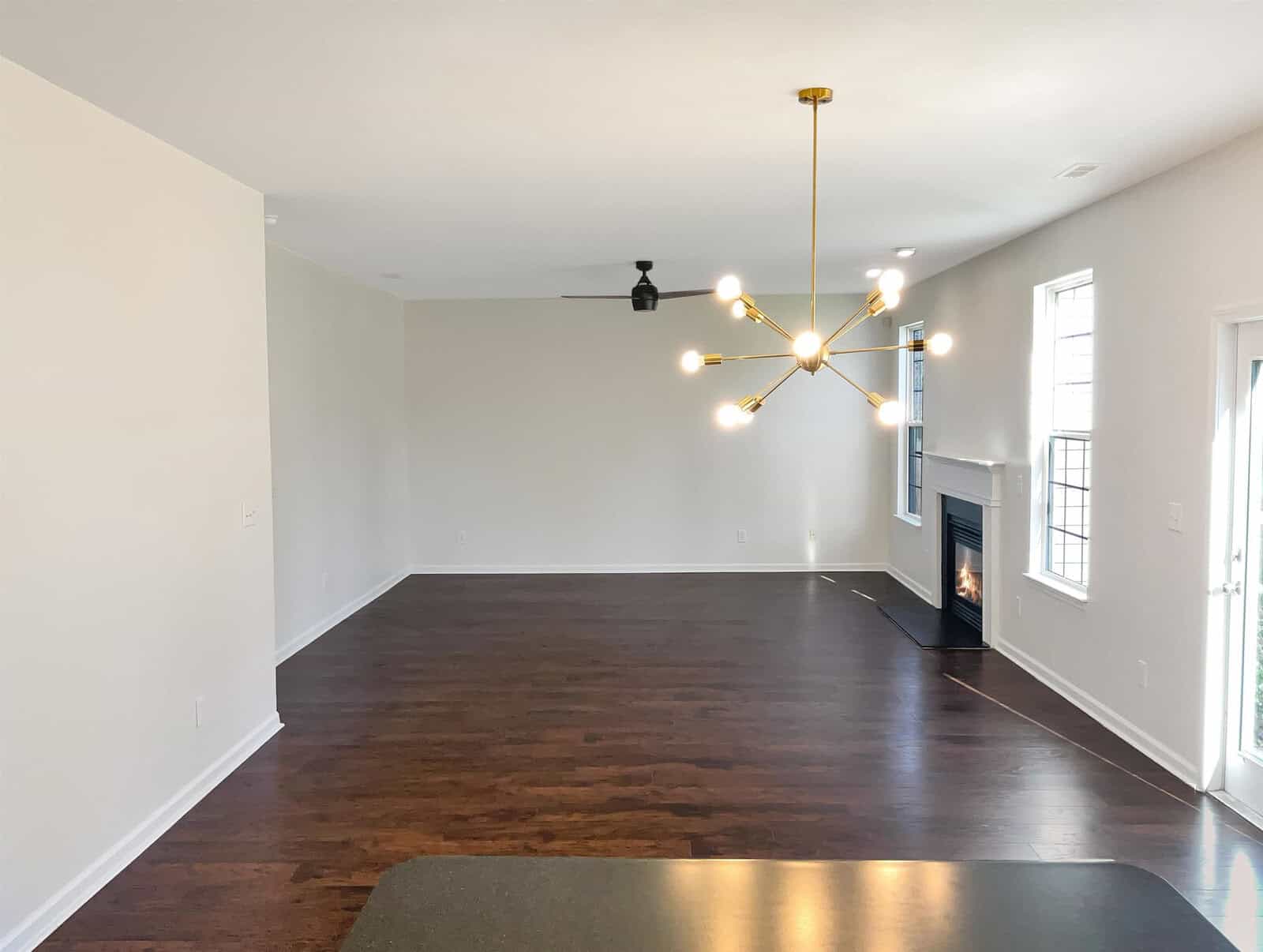 A modern, empty living room with dark hardwood floors and white walls. Features a sputnik chandelier, a ceiling fan, and a fireplace next to tall windows. Natural light enters through the windows and a glass door adjacent to the fireplace.