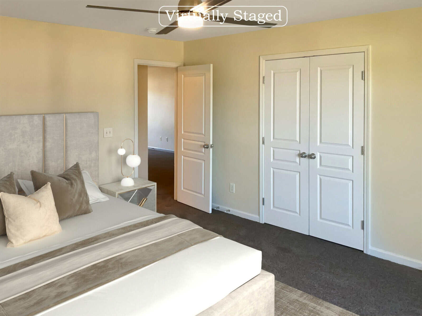 A virtually staged bedroom with cream walls features a modern bed with neutral bedding, a stylish side table with a lamp, and a ceiling fan. The room has a closed double door closet and an open door leading to another room.