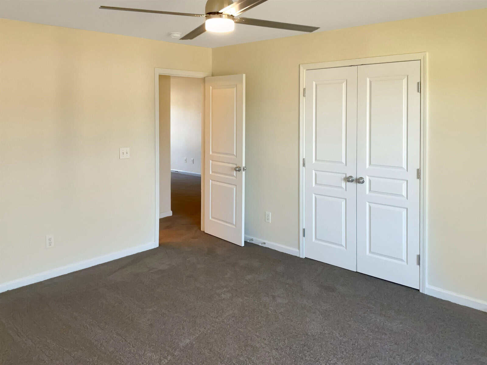 An empty room with beige walls and carpeted floor. A ceiling fan is mounted above. There are two doors: a closed double-door closet and an open door leading to another room.