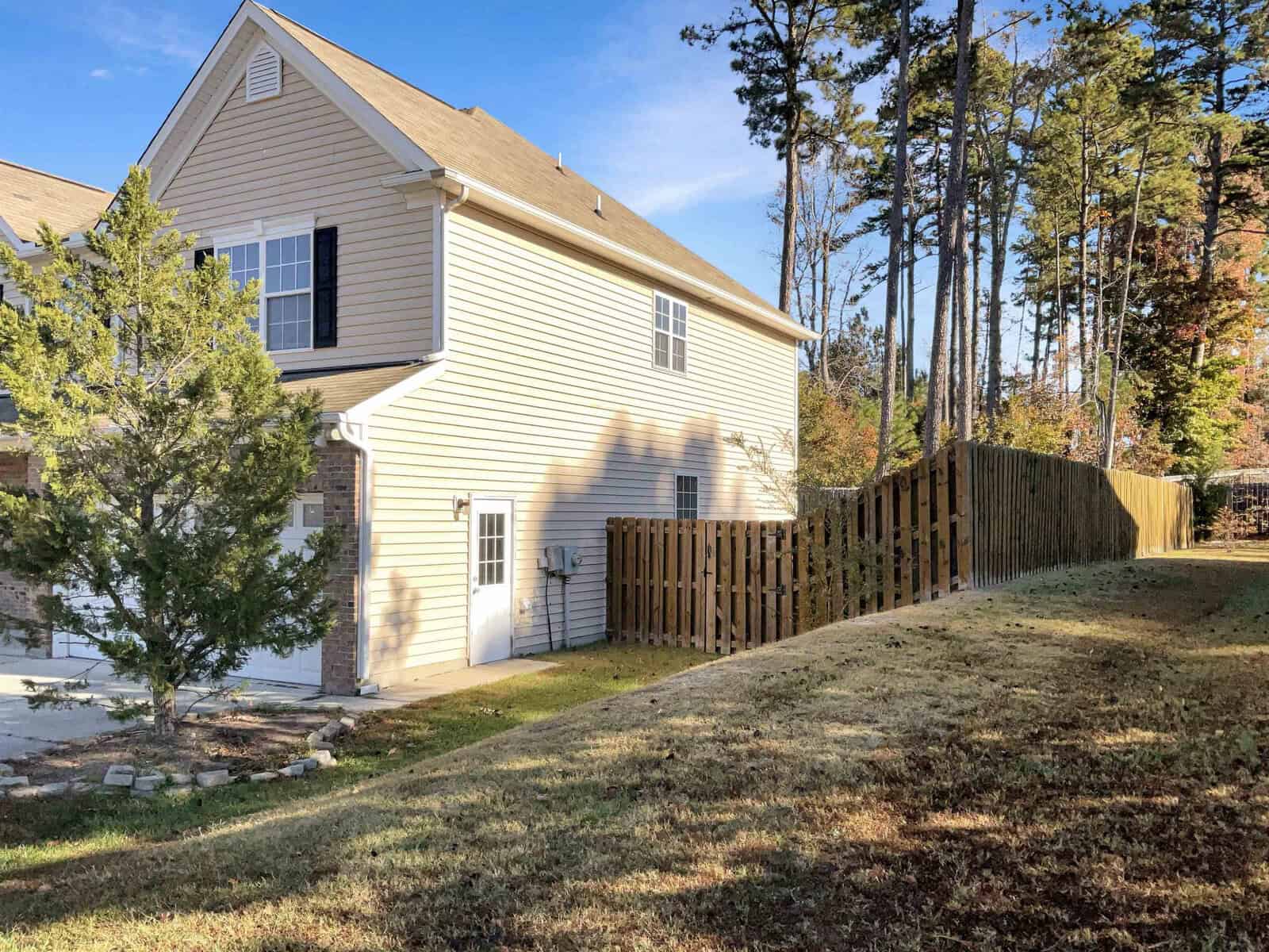 A two-story beige house with a sloped roof sits beside a wooden fence. A driveway is visible, and the yard is bordered by tall trees. It's a sunny day with a clear blue sky.