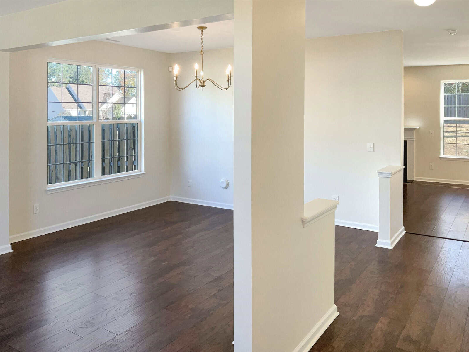 An empty, unfurnished room with dark hardwood floors and light-colored walls. A chandelier hangs from the ceiling. Large windows allow natural light to fill the space, and part of an adjoining room is visible.