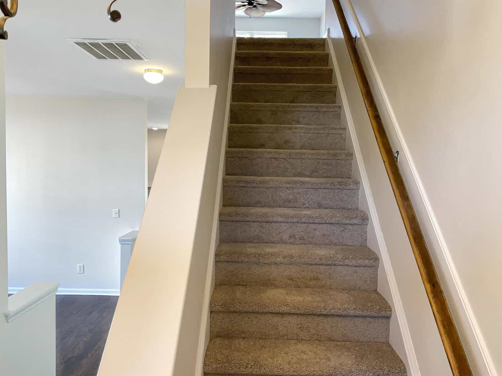 A carpeted staircase with a wooden handrail leads upstairs in a well-lit interior. The walls are painted in a light color, and a ceiling light fixture is visible at the top of the stairs.