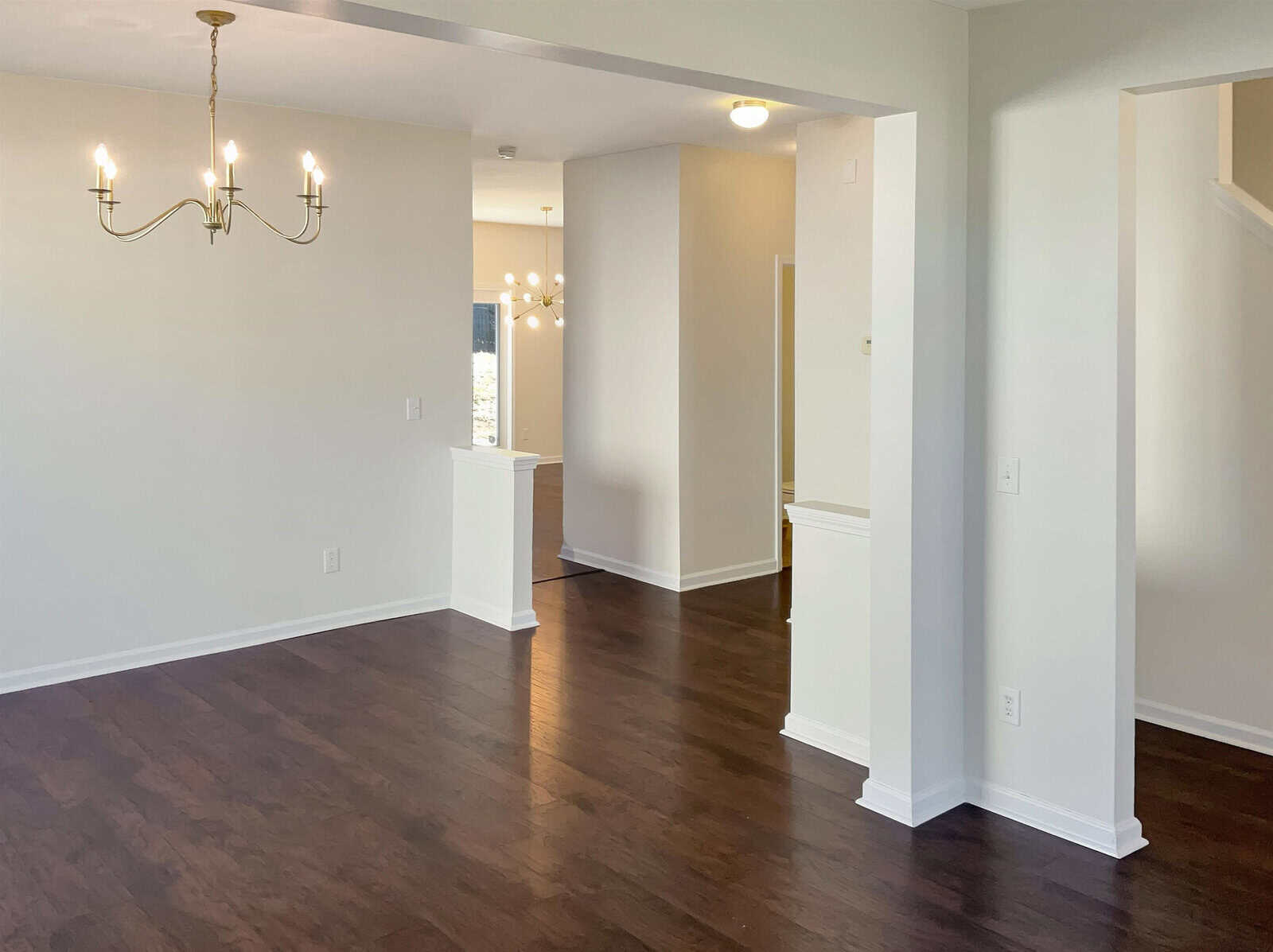 A spacious, empty dining room features dark hardwood floors, white walls, and a modern chandelier. The room opens into a hallway with additional lighting, creating a bright and open feel.