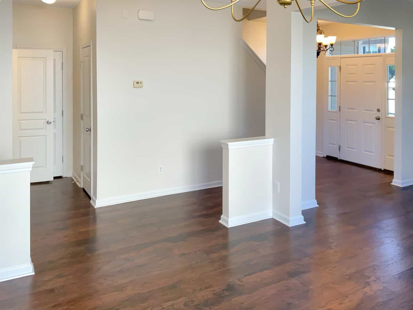 A bright, empty room with polished wooden floors and white walls, featuring a staircase, two doors, and a chandelier. Sunlight streams through the windows, illuminating the space.