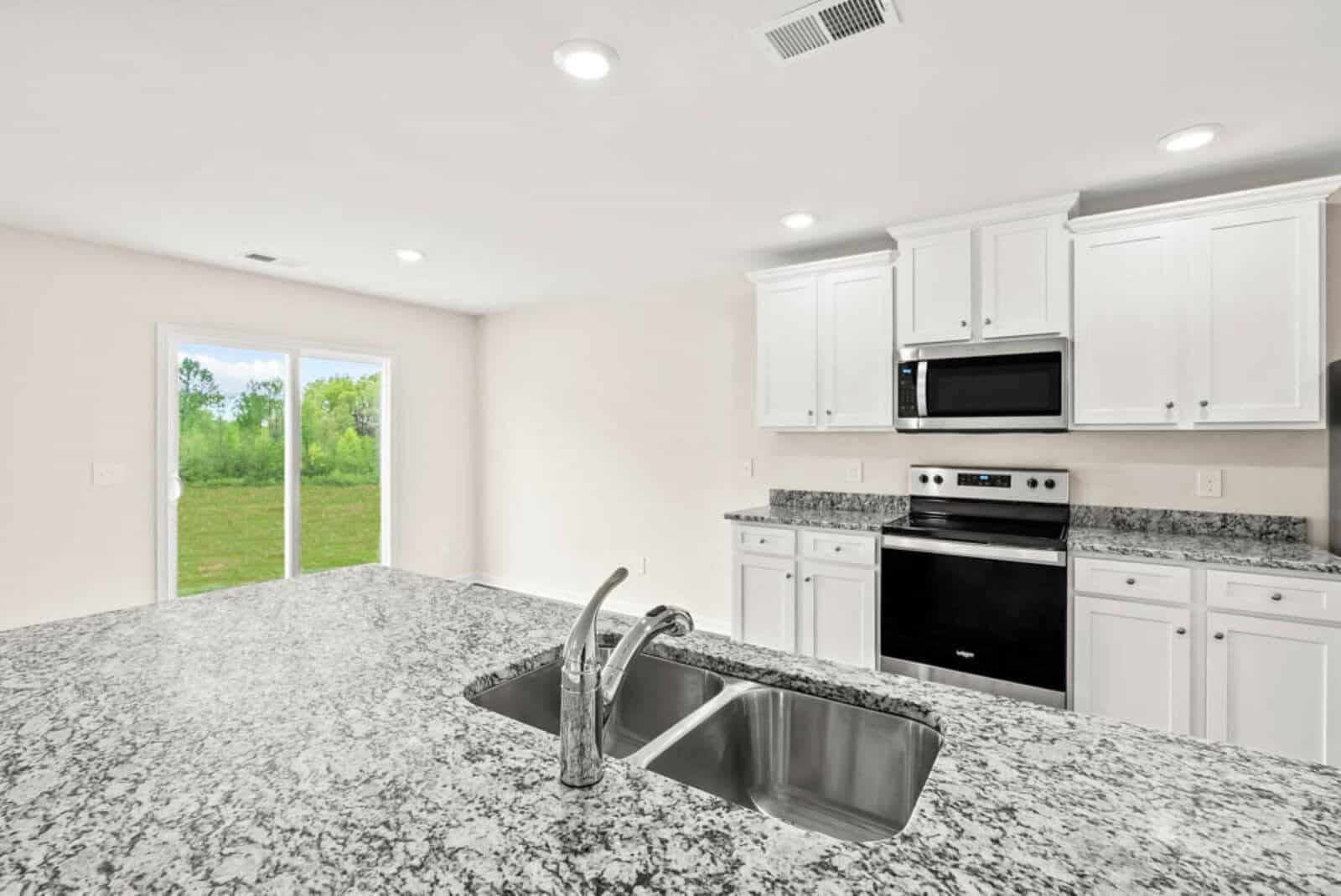 A modern kitchen with granite countertops, a double sink, and stainless steel appliances, including a stove and microwave. White cabinets line the walls, and a sliding glass door leads to a green outdoor area.