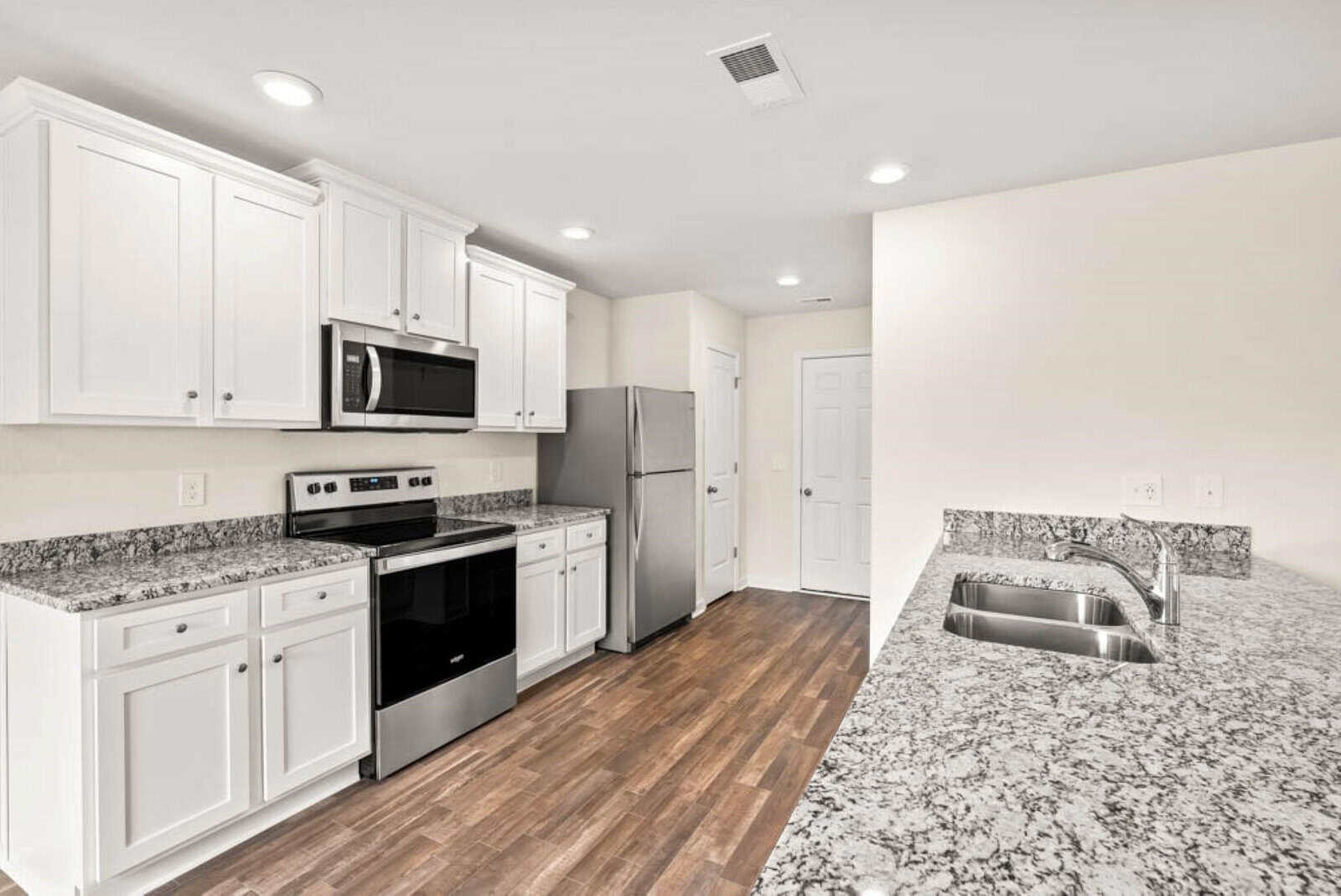 Modern kitchen with white cabinets, stainless steel appliances, and granite countertops. The room features a stove, microwave, refrigerator, and a dual sink set in an island counter. The floor is made of polished wood. Bright ceiling lights illuminate the space.
