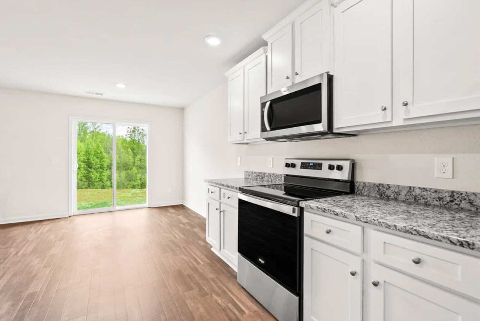A modern kitchen with white cabinets and granite countertops. There is a stainless steel oven with a matching microwave above it. Wood flooring extends into an adjacent room with large glass doors opening to a green outdoor area.