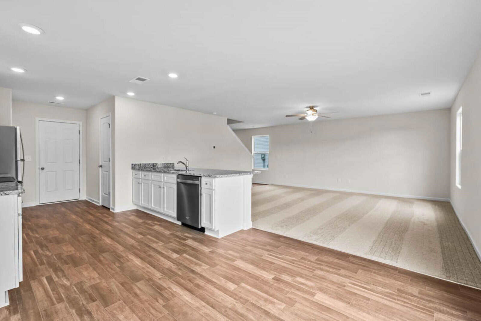 Open-concept living space with a kitchen featuring white cabinets, granite countertops, and stainless steel appliances. Adjacent is a carpeted area with a ceiling fan and natural light from a window. Wood flooring is visible in the kitchen area.