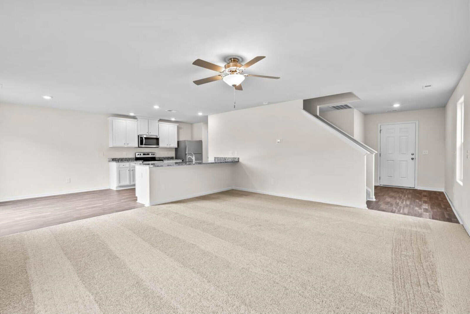 Modern living area with beige carpet, a ceiling fan, and white walls. Open kitchen with white cabinets, stainless steel appliances, and a tile backsplash. Wooden staircase and dark hardwood flooring near the entrance.