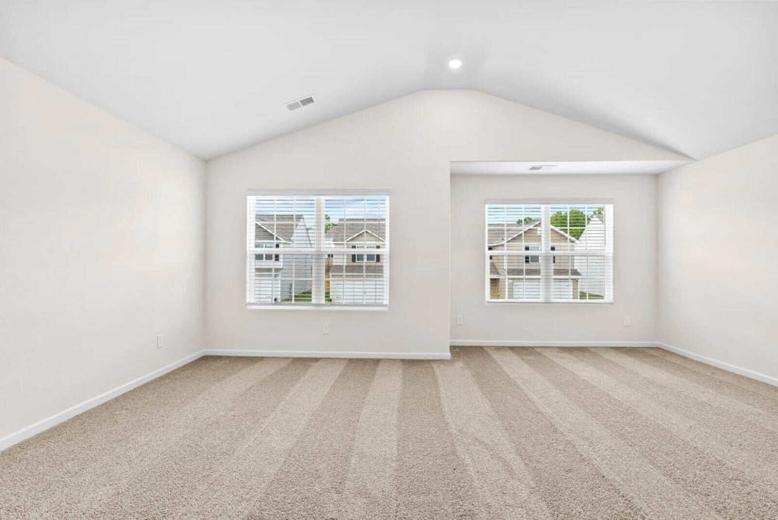 An empty, carpeted room with vaulted ceilings and two large windows that provide natural light. The walls are painted white, and there's a small alcove near one side. The view outside shows neighboring houses.