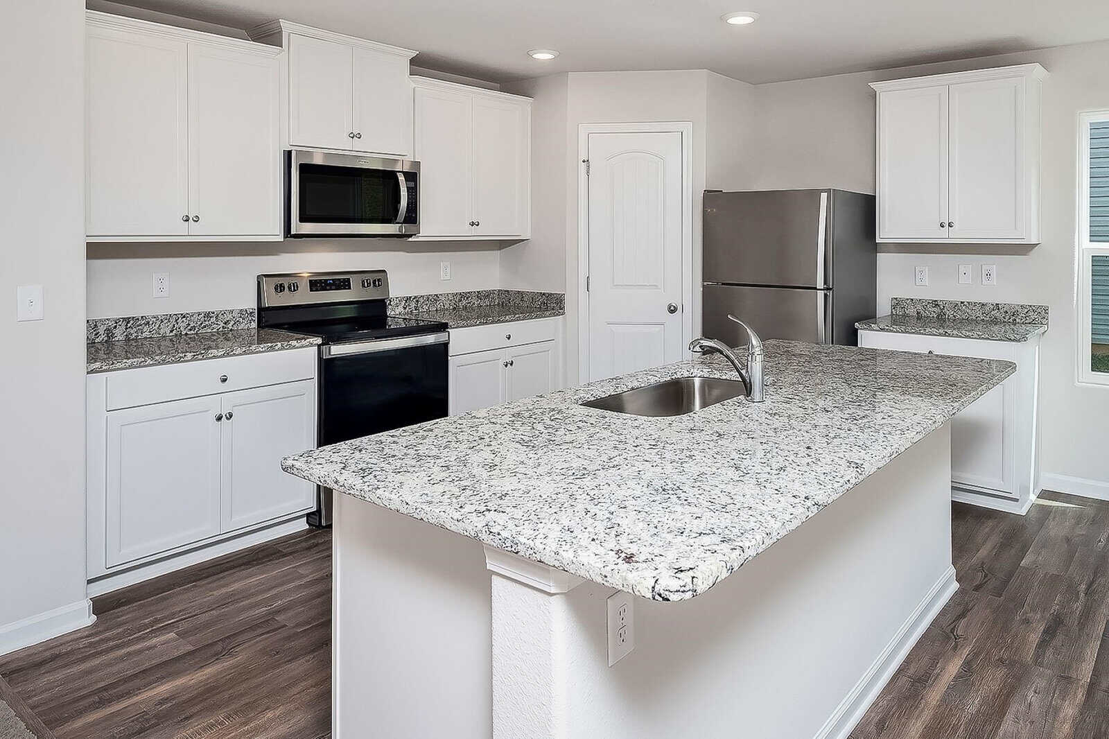 Modern kitchen with white cabinets, stainless steel appliances, and a large granite island with a built-in sink. Dark wood flooring contrasts with the light cabinetry. A door and windows are visible.