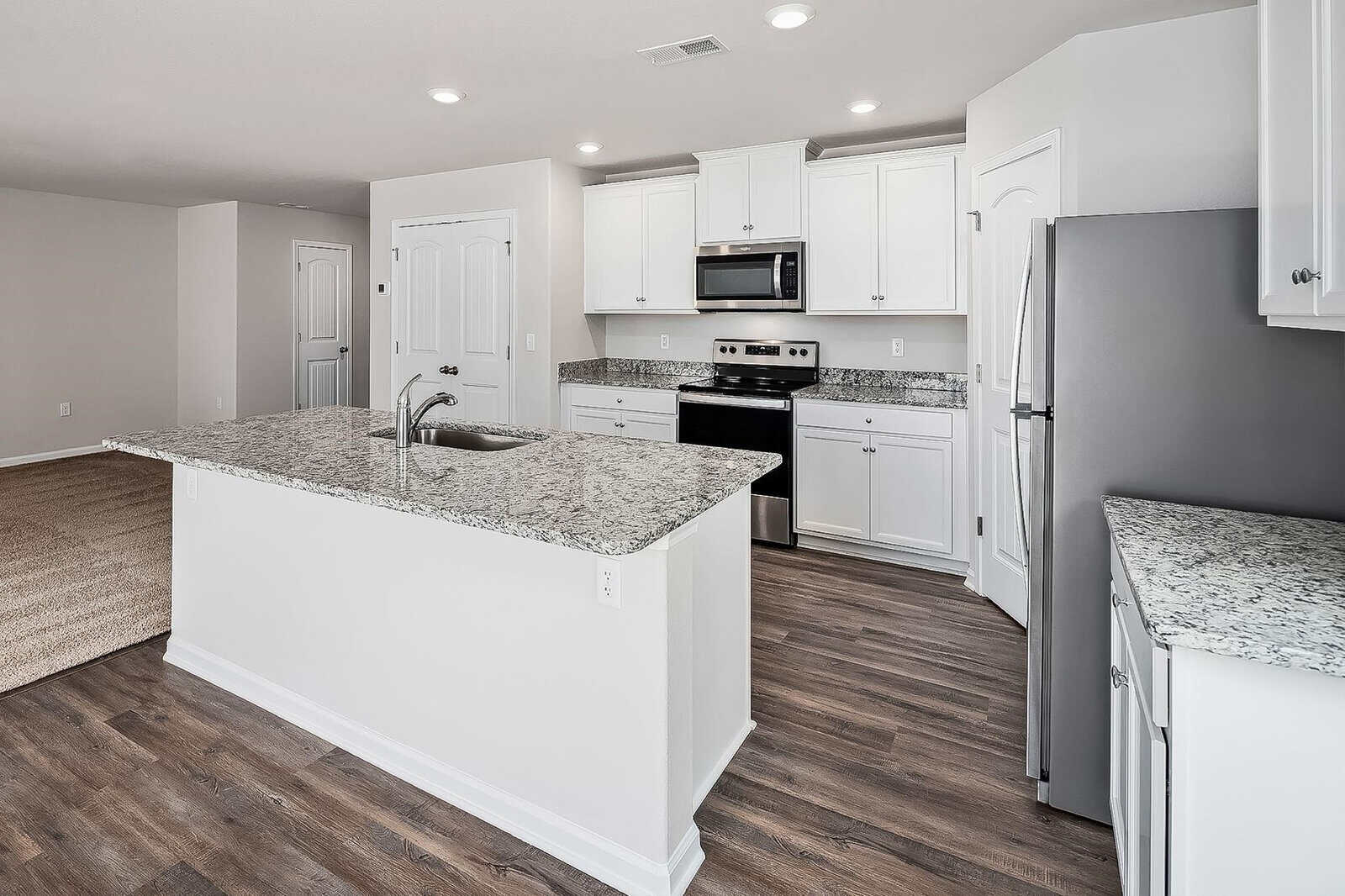 A modern kitchen with white cabinets, stainless steel appliances, and granite countertops. A large island with a sink is centrally located. The room features hardwood flooring and recessed lighting, with an adjacent open living space.