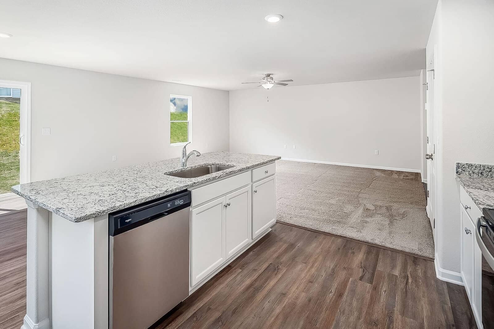 A modern, open-concept kitchen and living area with light wood flooring. The kitchen features a large granite island with a sink and dishwasher. Carpeting begins just beyond the kitchen, and natural light enters through windows and a glass door.